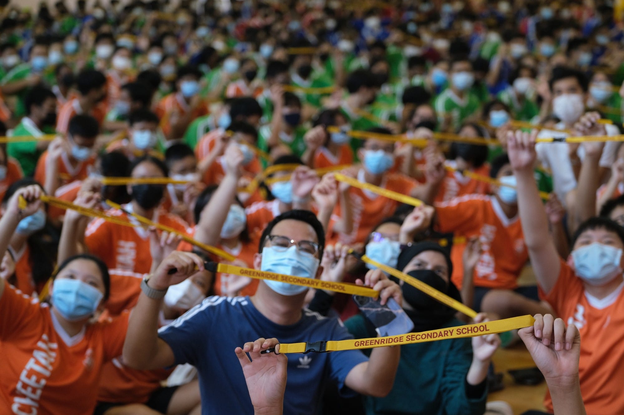 Large masked crowd holds up yellow Greenridge Secondary School lanyards in an indoor assembly hall.